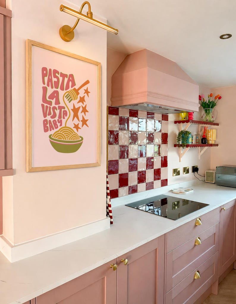 May include: A pink kitchen with a framed poster that says "Pasta La Vista Baby". The kitchen features pink cabinets, a white countertop, and a red and white checkered backsplash. A gold light fixture is mounted above the poster.