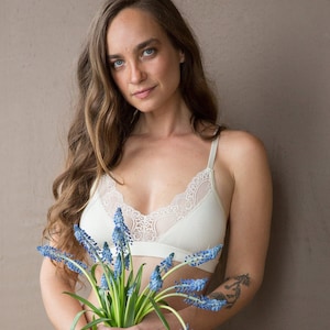 Op de afbeelding: Een vrouw draagt een witte kanten bralette en bijpassende ondergoed. Ze houdt een boeket blauwe bloemen vast.