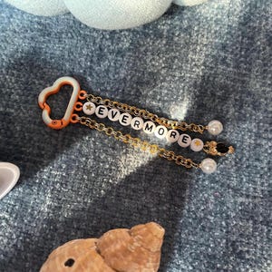 May include: A keychain with the word "EVERMORE" spelled out in white letter beads. The keychain has gold chains, a heart-shaped orange clasp, and pearl and black bead accents. The background is a textured blue surface.