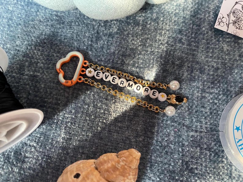 May include: A keychain with the word "EVERMORE" spelled out in white letter beads. The keychain has gold chains, a heart-shaped orange clasp, and pearl and black bead accents. The background is a textured blue surface.