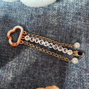 May include: A keychain with the word "EVERMORE" spelled out in white letter beads. The keychain has a gold chain, a white and orange clasp, and pearl and star accents.