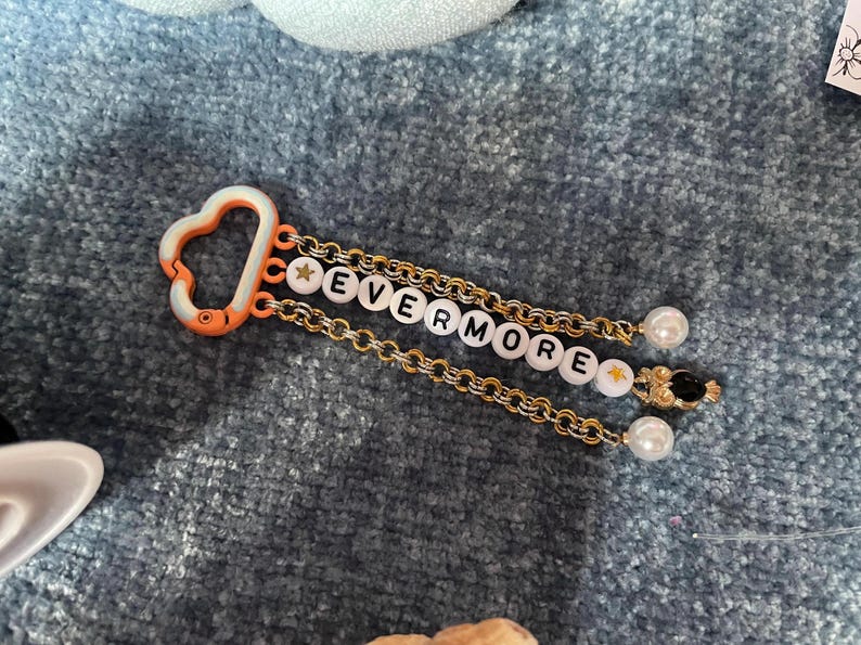 May include: A keychain with the word "EVERMORE" spelled out in white letter beads. The keychain has a gold chain, a white and orange clasp, and pearl and star accents.