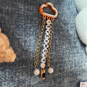 May include: A decorative charm featuring a cloud-shaped orange and white top piece. Gold chains hang below, with white letter beads spelling "EVERMORE". The charm includes a gold star bead, a small owl charm, and two pearl-like beads.