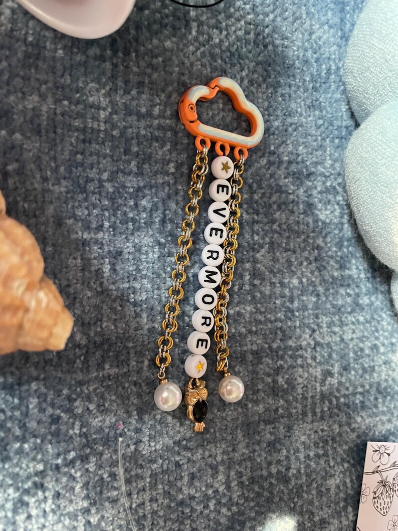 May include: A decorative charm featuring a cloud-shaped orange and white top piece. Gold chains hang below, with white letter beads spelling "EVERMORE". The charm includes a gold star bead, a small owl charm, and two pearl-like beads.