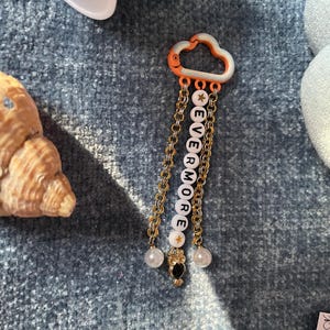 May include: A cloud-shaped keychain with the word "EVERMORE" spelled out in white letter beads. The keychain has gold-colored chains, star-shaped beads, and pearl accents. An owl charm and orange accents complete the design.
