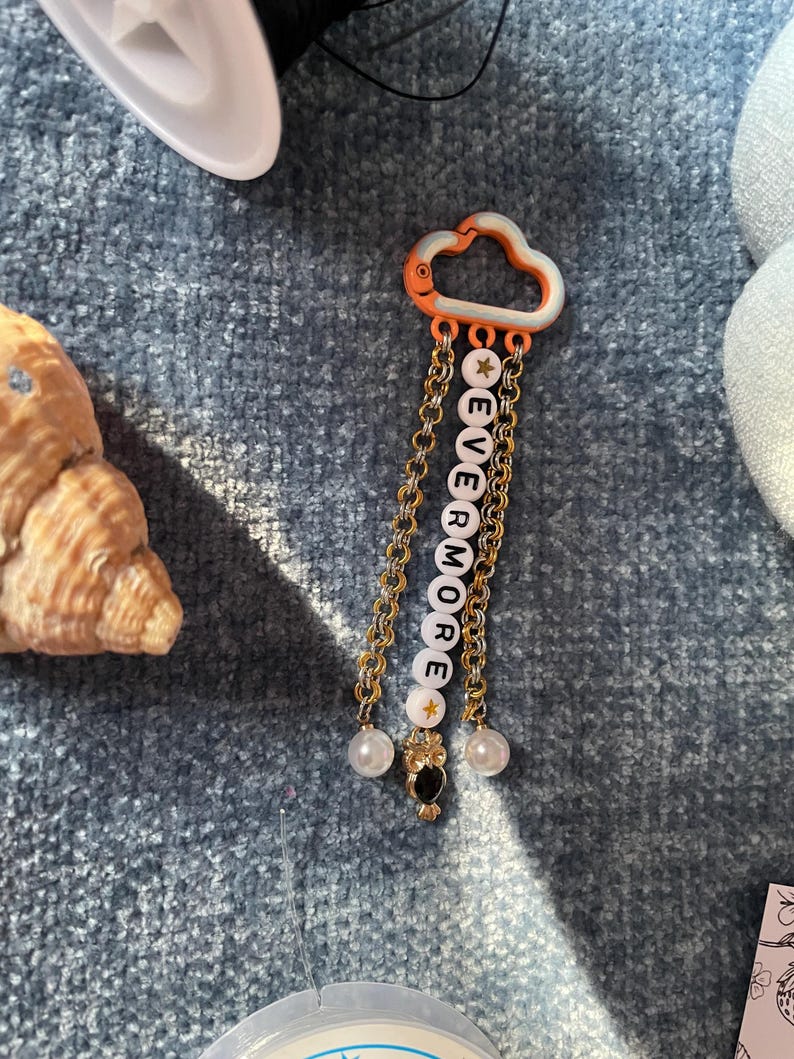 May include: A cloud-shaped keychain with the word "EVERMORE" spelled out in white letter beads. The keychain has gold-colored chains, star-shaped beads, and pearl accents. An owl charm and orange accents complete the design.