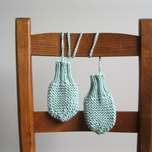 May include: Two knitted baby mittens in a light green colour hanging on a wooden chair back.