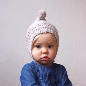 May include: A baby wearing a light pink knitted hat with a pom-pom on top. The hat has a flap that covers the ears and neck. The baby is wearing a blue denim shirt.