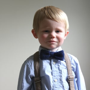 May include: A young boy wearing a light blue button-down shirt with a dark blue bow tie and brown braces.