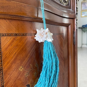 May include: A decorative seashell tassel with a light blue cord. The tassel is made of thick, twisted yarn. The seashell is white and attached to the cord above the tassel. The tassel hangs in front of a wooden cabinet.