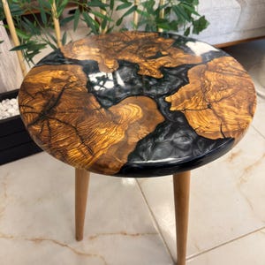 Puede incluir: Mesa auxiliar redonda con una tapa de madera con un patrón de vetas único y detalles de resina negra. La mesa tiene tres patas de madera. La parte superior tiene aproximadamente 61 cm de diámetro.