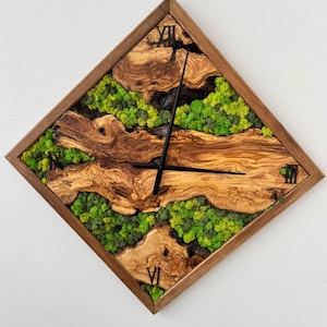 Op de afbeelding: Een ruitvormige houten wandklok met een natuurlijke houtnerf afwerking. De wijzerplaat heeft een combinatie van hout, groen mos en zwarte wijzers. Romeinse cijfers markeren de uren.