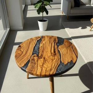 May include: Round wooden side table with a unique design. The tabletop features a natural wood grain pattern with dark gray resin accents. The table has three slender, light-colored wooden legs. The table is approximately 24 inches in diameter.