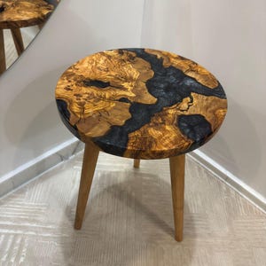 May include: A round side table with a wooden top and three wooden legs. The tabletop features a unique design with a combination of light brown wood and dark gray resin. The table is approximately 18 inches in diameter.