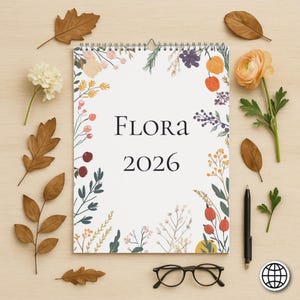 May include: A spiral-bound calendar with the text "FLORA 2026" surrounded by floral illustrations. The image includes a pen, eyeglasses, and decorative leaves and flowers, all arranged on a light wooden surface.