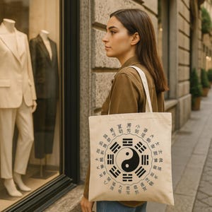 Op de afbeelding: Een beige draagtas met een witte band, met een zwart Yin en Yang symbool omringd door Chinese karakters. De tas wordt gedragen door een persoon die een bruine jas draagt.