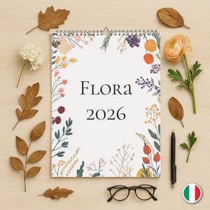 Puede incluir: Un calendario en espiral con la palabra "FLORA" y el año "2026" en negro. El calendario está decorado con ilustraciones florales y hojas secas. También hay un bolígrafo negro, gafas y una flor.