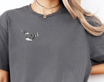 May include: A charcoal gray t-shirt with a small embroidered design of two orcas swimming. The shirt is worn by a person with long blonde hair and blue jeans.