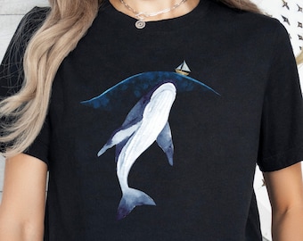 May include: Black t-shirt featuring a watercolor illustration of a whale swimming underwater with a small sailboat on the surface. The whale is white with blue accents, and the water is a deep blue. The shirt has short sleeves.