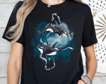 May include: Black t-shirt with a watercolor design of two orcas swimming in a blue and gray ocean. The orcas are black and white with a white patch on their bellies.