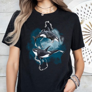 May include: Black t-shirt with a watercolor design of two orcas swimming in a blue and gray ocean. The orcas are black and white with a white patch on their bellies.