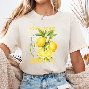 May include: Cream-colored t-shirt featuring a watercolor illustration of lemons with green leaves and the text "easy peasy lemon squeezy" in yellow. The shirt is made of soft material and is suitable for casual wear.
