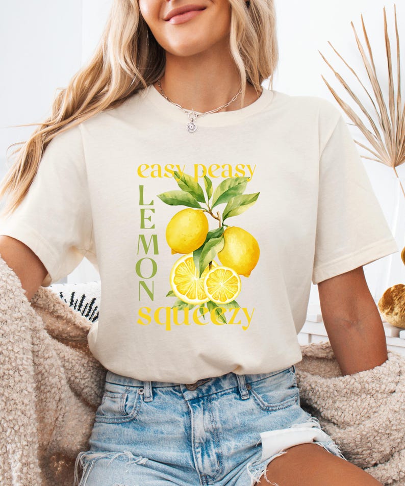 May include: Cream-colored t-shirt featuring a watercolor illustration of lemons with green leaves and the text "easy peasy lemon squeezy" in yellow. The shirt is made of soft material and is suitable for casual wear.