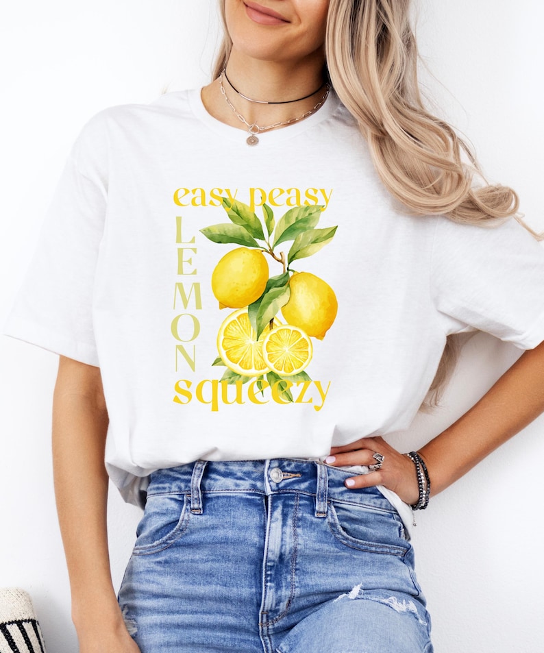 May include: White t-shirt featuring a watercolor illustration of lemons with green leaves. The text "easy peasy lemon squeezy" is printed in yellow. The shirt is paired with blue jeans.