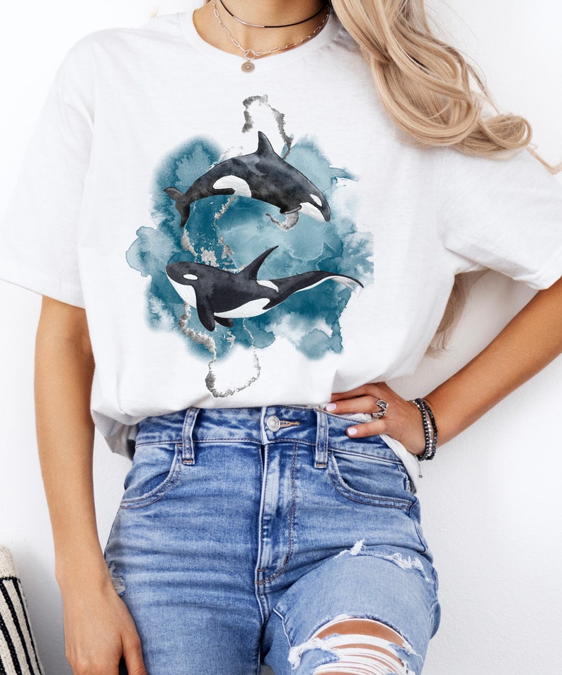 May include: White t-shirt with a watercolor design of two orcas swimming in blue water.