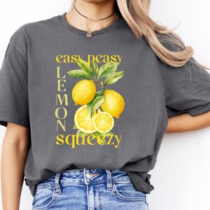 May include: A dark gray t-shirt featuring a yellow lemon graphic with the words "easy peasy lemon squeezy". The graphic includes yellow lemons and green leaves. The shirt is a casual style.