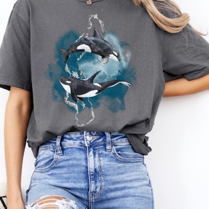 May include: A charcoal grey t-shirt with a watercolor design of two orcas swimming in a blue and white ocean. The orcas are black and white with white accents.