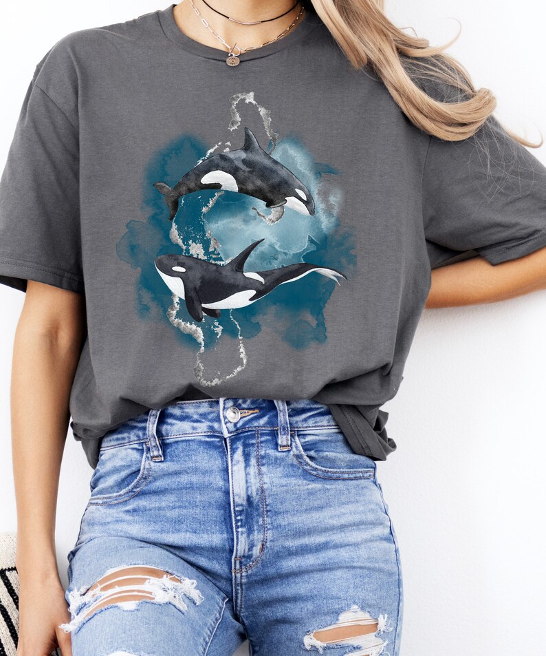 May include: A charcoal grey t-shirt with a watercolor design of two orcas swimming in a blue and white ocean. The orcas are black and white with white accents.
