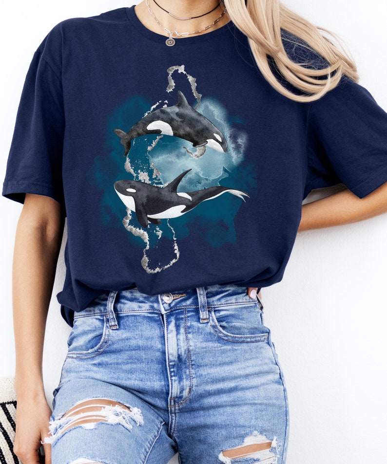 May include: A navy blue t-shirt with a watercolor design of two orcas swimming in a blue and white ocean. The orcas are black and white and have a realistic design.