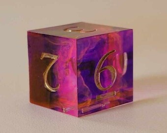 May include: A purple and pink resin dice with gold numbers. The dice shows the number 6.
