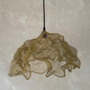 May include: A pendant light fixture featuring a gold-toned, fine wire mesh shade with an organic, ruffled, and undulating shape. The shade is suspended from a thin black cord and houses a single light bulb visible through the metallic netting.