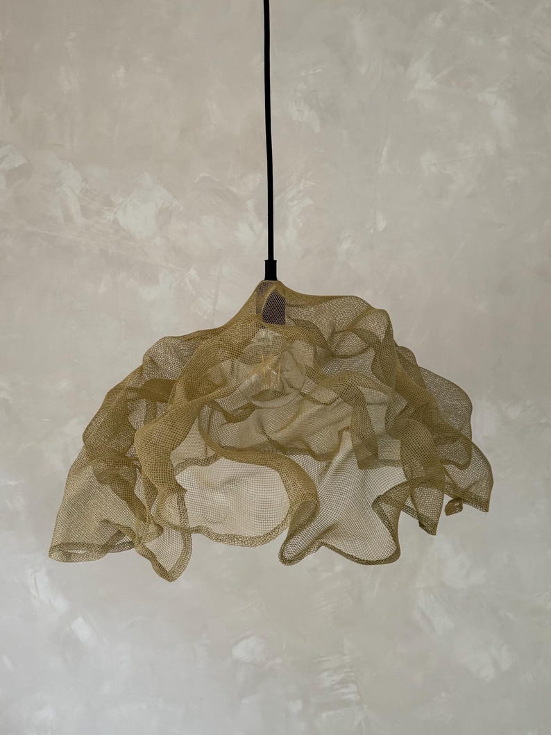 May include: A pendant light fixture featuring a gold-toned, fine wire mesh shade with an organic, ruffled, and undulating shape. The shade is suspended from a thin black cord and houses a single light bulb visible through the metallic netting.