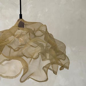 May include: A pendant light fixture featuring a gold-colored, fine wire mesh shade with an organic, undulating shape. The shade is suspended by a thin black cord and includes a single visible light bulb inside the metallic, semi-transparent structure.