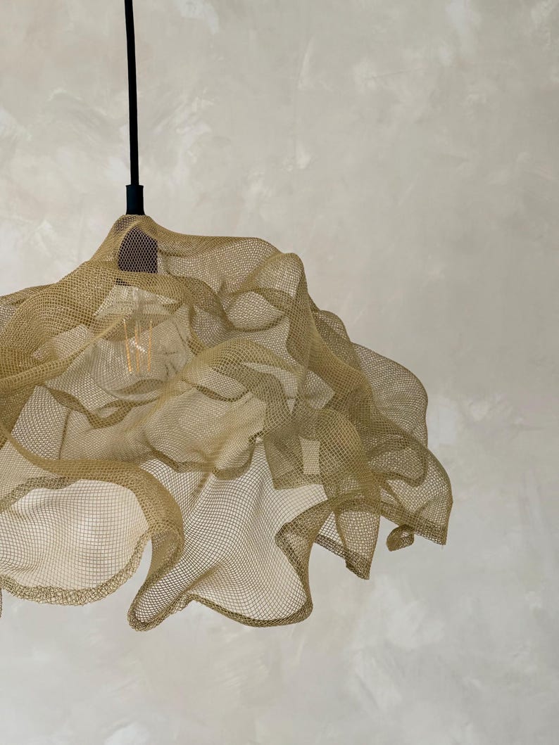 May include: A pendant light fixture featuring a gold-colored, fine wire mesh shade with an organic, undulating shape. The shade is suspended by a thin black cord and includes a single visible light bulb inside the metallic, semi-transparent structure.
