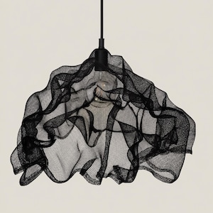 Può includere: Una lampada a sospensione nera caratterizzata da un paralume in rete drappeggiata e scultorea, con una forma irregolare e fluida. Il paralume è sospeso a un cavo e un portalampada neri, con una lampadina visibile attraverso la struttura a griglia.