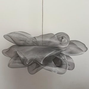 Könnte beinhalten: Eine moderne, abstrakte Leuchte. Sie besteht aus einem grauen, netzartigen Material und hängt an einem dünnen Kabel. Das Design zeigt geschichtete, fließende Formen, die ein weiches, wolkenartiges Aussehen erzeugen. Der Hintergrund ist schlicht weiß.