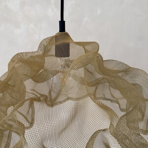 May include: A pendant light fixture featuring a gold-colored, fine wire mesh shade with a wavy, organic form. The shade is suspended from a black cord and a black cylindrical socket, which holds a single, visible light bulb inside the mesh structure.