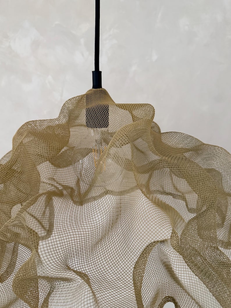 May include: A pendant light fixture featuring a gold-colored, fine wire mesh shade with a wavy, organic form. The shade is suspended from a black cord and a black cylindrical socket, which holds a single, visible light bulb inside the mesh structure.
