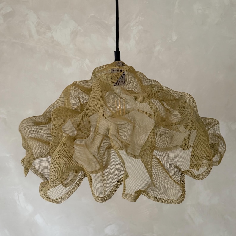 May include: A pendant light fixture featuring a gold-colored, fine wire mesh shade with a fluid, rippled, and layered structure. The shade is suspended from a thin black cord and contains a single visible light bulb in the center.