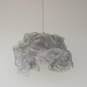 May include: A gray, cloud-like pendant light fixture. The shade is made of a sheer, ruffled material, suspended from a thin cord and a brass-colored ceiling mount. The light fixture is a modern design element.