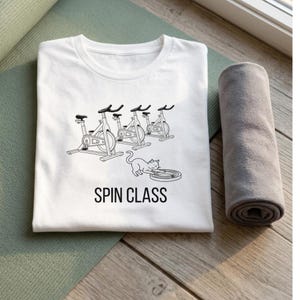 May include: White t-shirt with a black graphic of spin bikes and a cat playing with a record. The text "SPIN CLASS" is below the graphic. A rolled gray towel is next to the shirt.