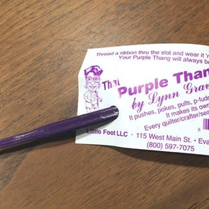 Könnte beinhalten: Purple Thang Nähwerkzeug von Lynn Graves. Es ist ein violettes Plastikwerkzeug mit einem Schlitz für ein Band. Das Werkzeug wird zum Drücken, Stochern, Ziehen, P-Fudges, P-Turns und P-Stults verwendet. Die Karte sagt "Jeder Quilter/Handwerker/Schneider braucht einen!"