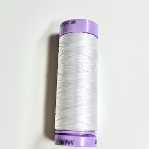Peut inclure: Une bobine de fil blanc avec un capuchon violet. L'étiquette indique "100% Coton". Le fil est bien enroulé autour de la bobine. La bobine est cylindrique et se dresse sur un fond blanc.