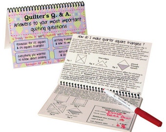 May include: A pink and white spiral bound book titled "Quilter's Q&A: Answers to your most important quilting questions". The book has a dry erase surface and includes instructions on how to make quarter square triangles. The book is open to a page with diagrams and instructions on how to make quarter square triangles.