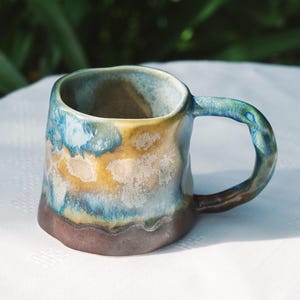 May include: A handmade ceramic mug with a unique, organic shape. The mug features a blend of colors, including blue, yellow, and brown, creating a textured, glazed finish. The handle is a matching color. This mug is perfect for coffee or tea.