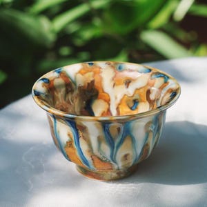 May include: A small, ceramic bowl with a unique, colorful glaze. The bowl features a mix of orange, brown, blue, and white hues, creating a marbled effect. The bowl is round with a slightly flared rim and a small, rounded base. The bowl is sitting on a white surface.
